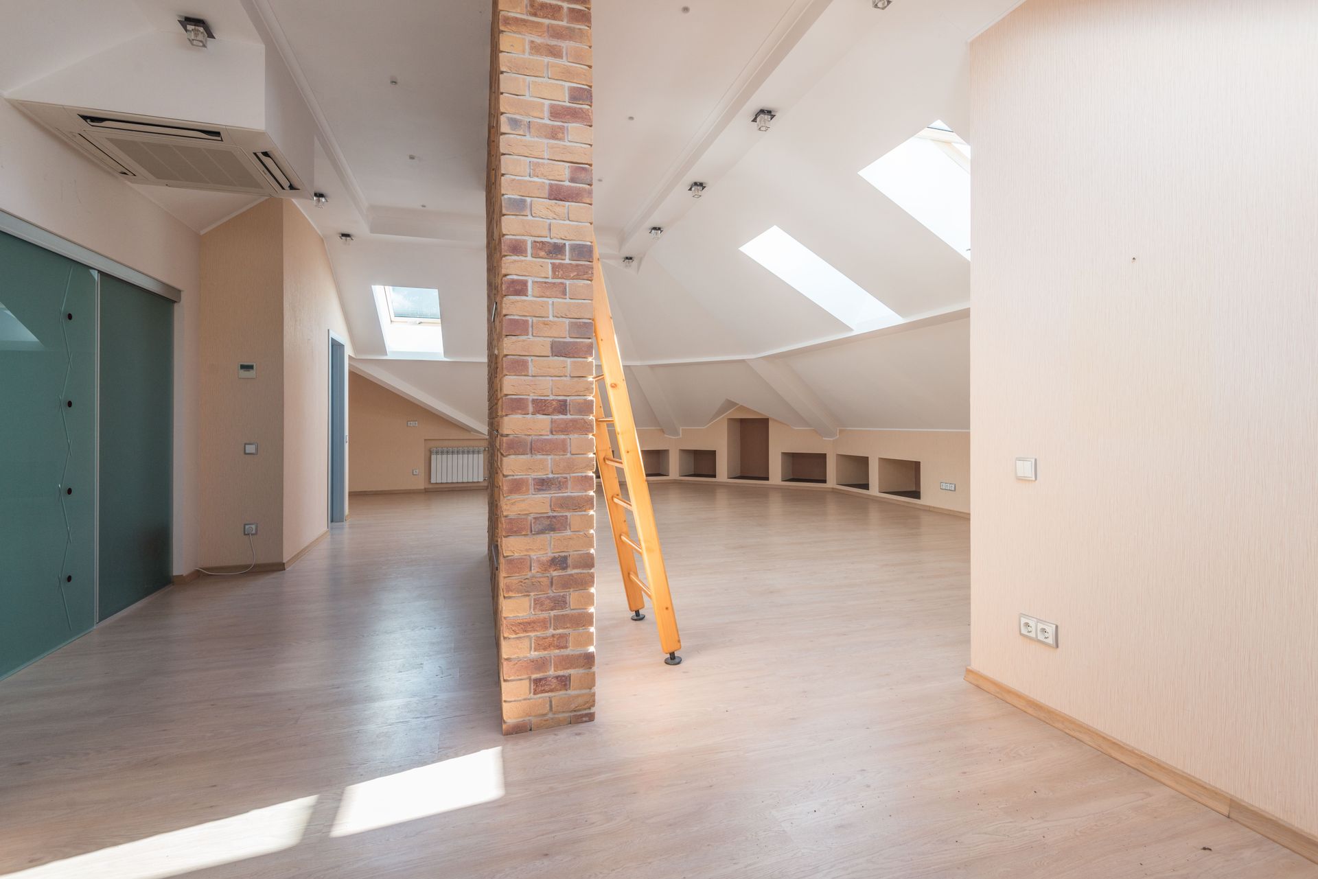Empty loft with brick column, skylights, light wood floors, and a leaning yellow ladder.