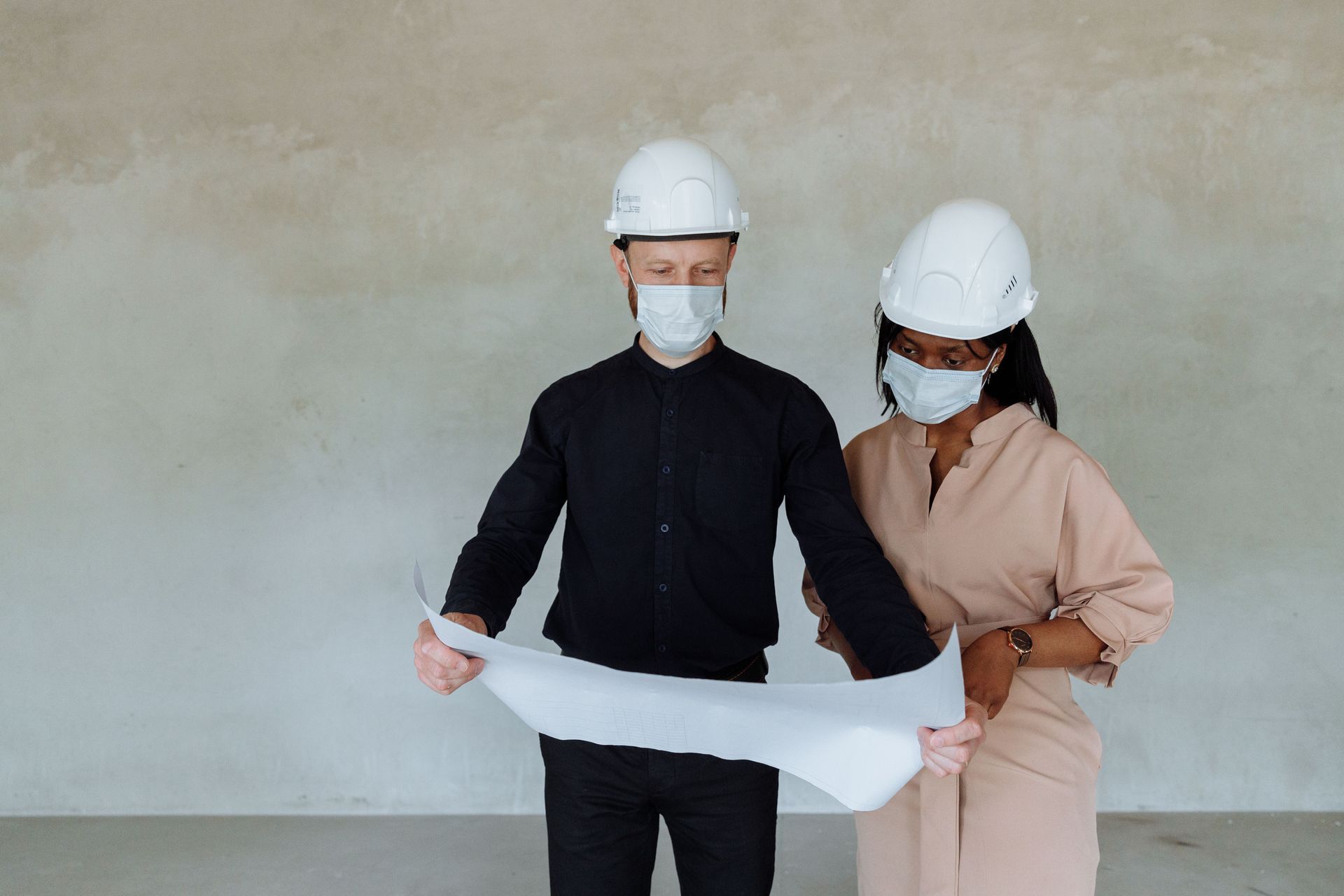 Two construction workers in hard hats and masks reviewing a blueprint indoors