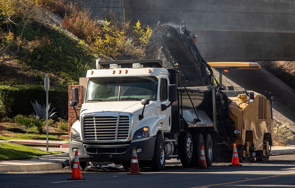 White dump truck and paving machine on a road with orange cones and steam rising nearby