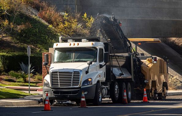 White dump truck and paving machine on a road with orange cones and steam rising nearby