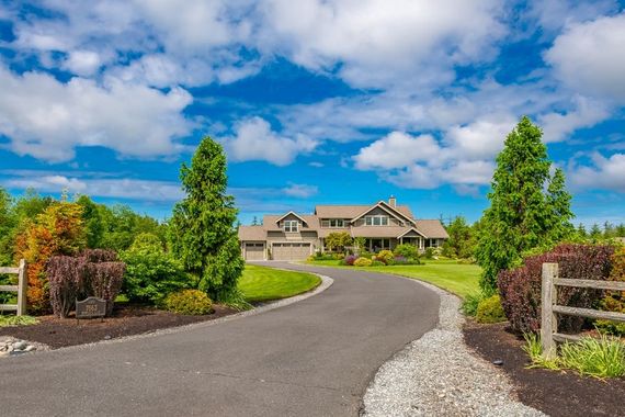 Curved driveway leading to a large house with manicured landscaping under a bright blue, cloudy sky