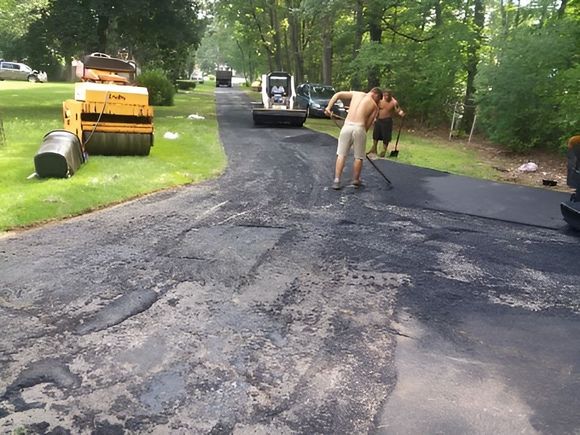 Road workers paving a cracked asphalt driveway beside parked construction equipment in a wooded area