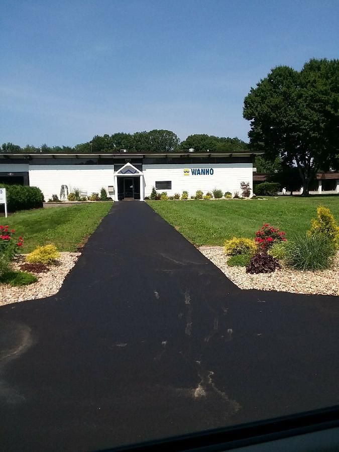 White building at the end of a black paved path, with lawns, flower beds, and trees on both sides.