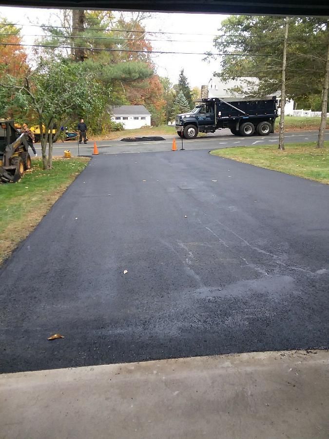 Freshly paved driveway leading to a street, with a black dump truck and orange cones in the distance.