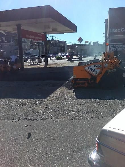 Orange roadwork machine parked beside a gas station on a sunny street.