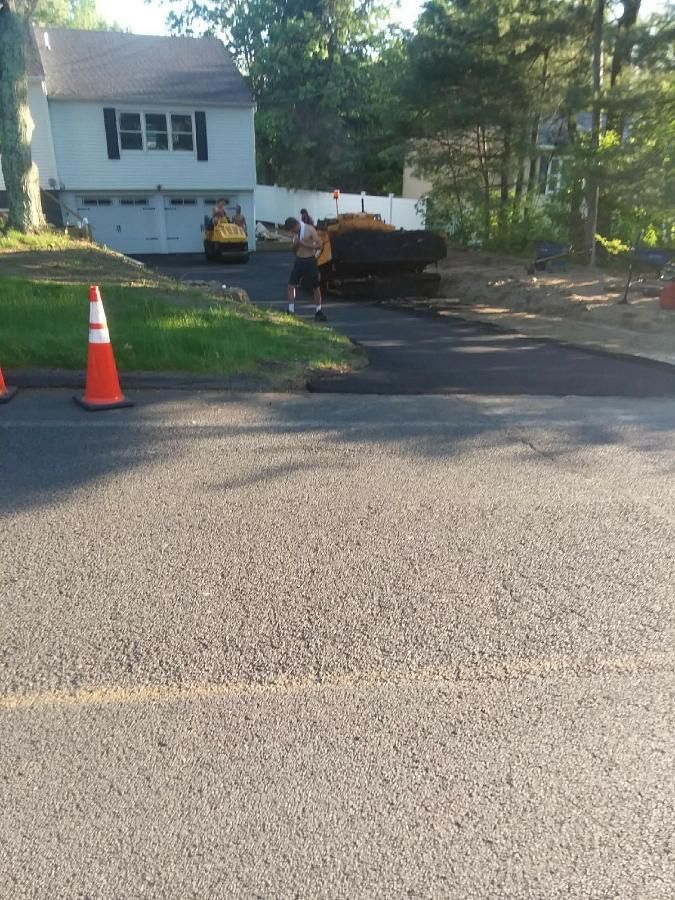 Driveway with an orange traffic cone, a person near a dark dumpster, and a white house in the background.