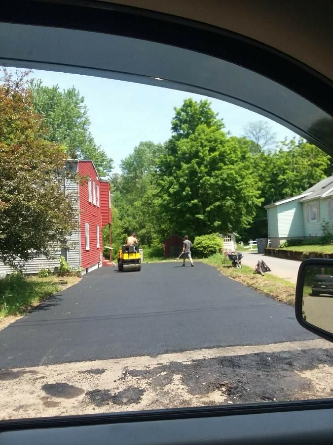 A paved neighborhood street with a person on a lawn tractor, another walking, and houses with trees nearby.
