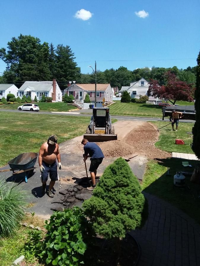 Two workers paving a backyard patio with black stones, surrounded by grass and suburban houses.
