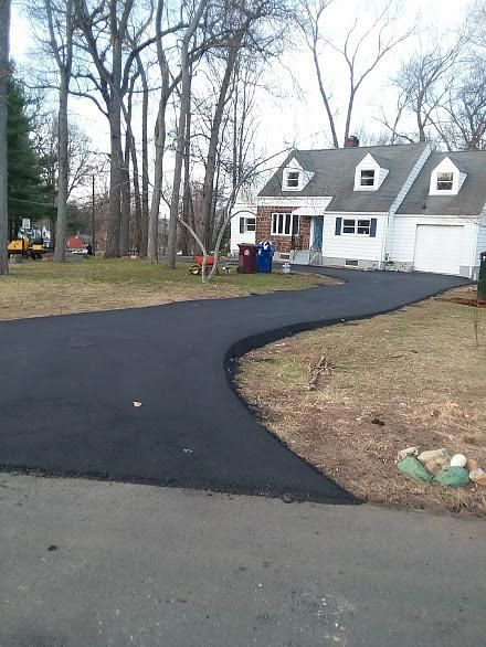 Freshly paved black driveway curving to a white house with garages and bare trees
