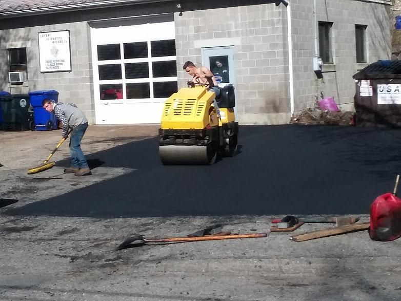 Workers paving a driveway beside a garage, with a yellow road roller on fresh black asphalt