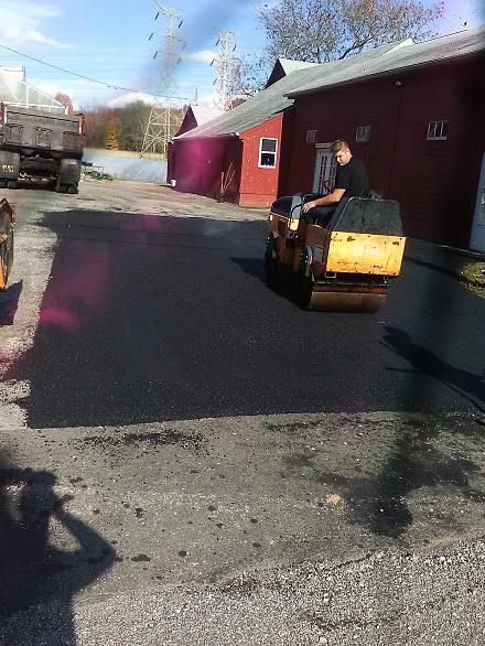 Person driving a small loader on a paved road beside a red building on a sunny day