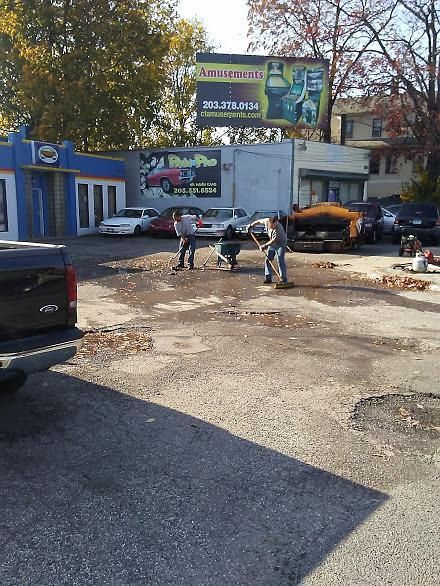 Workers fixing a pothole in a rough parking lot beside a blue building and billboard.