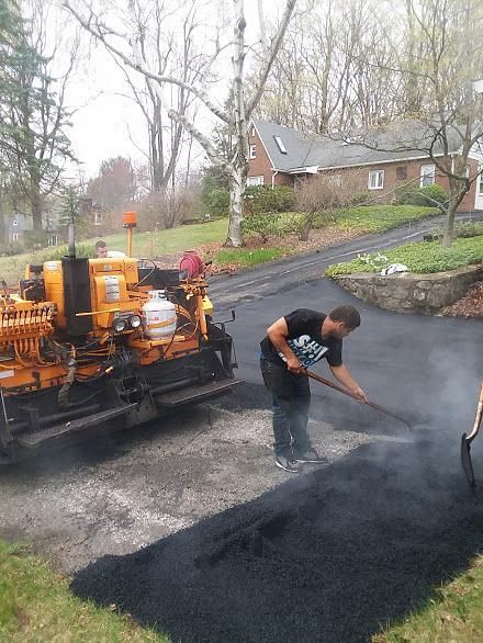 Worker spreading black asphalt beside paving machine on a wet residential street