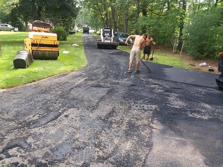 Workers paving a residential driveway with fresh black asphalt, yellow rollers and equipment nearby