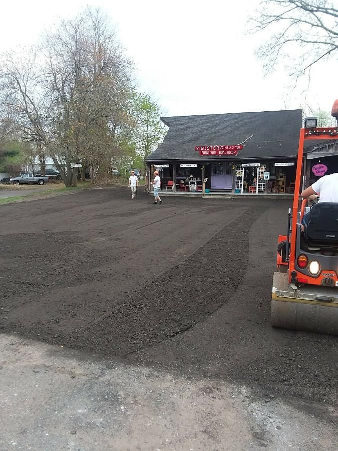 Roadwork crew paving a parking lot outside a storefront, with an orange roller in the foreground.