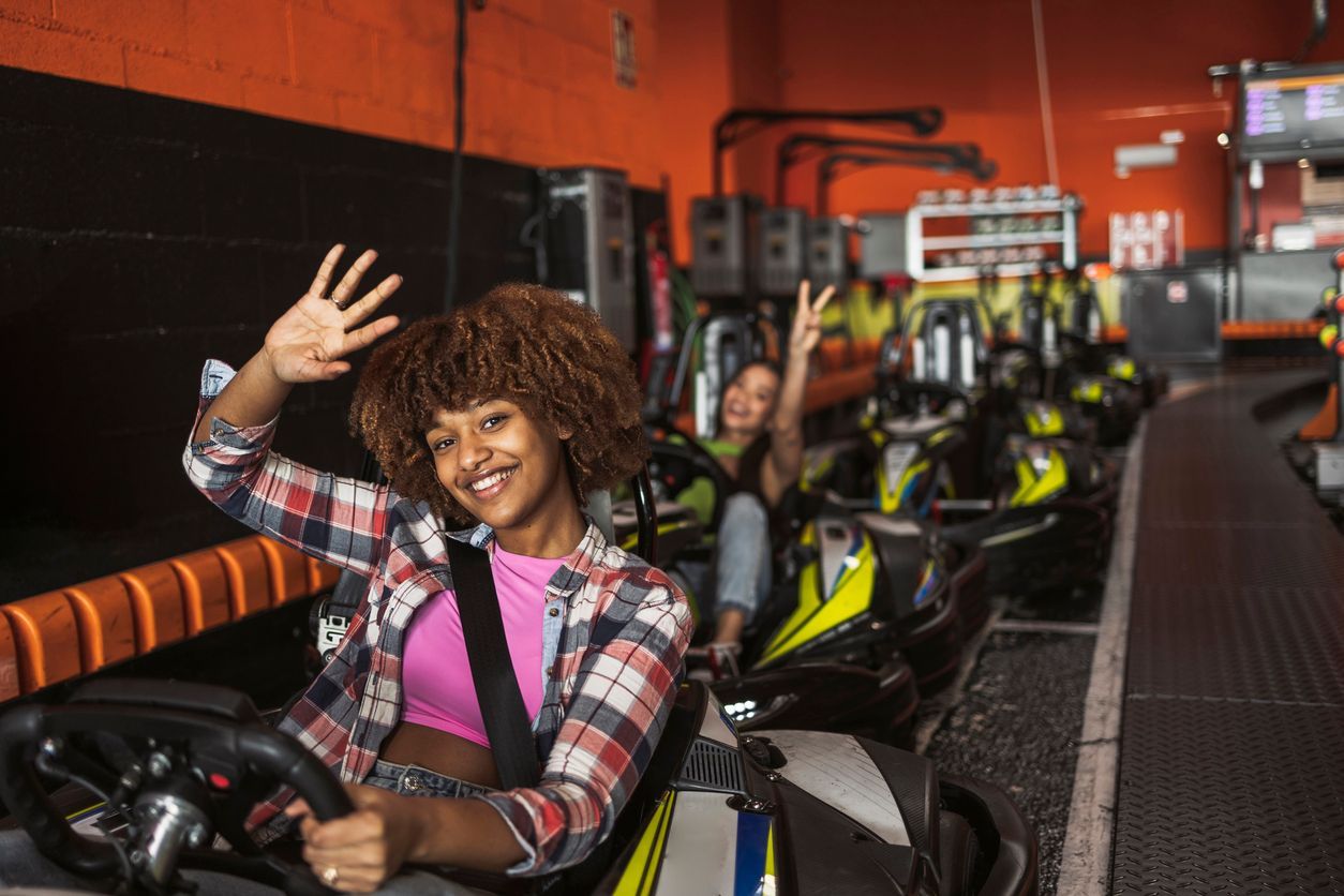 Two young women in go-karts waving, smiling. Indoor track, black and yellow karts.