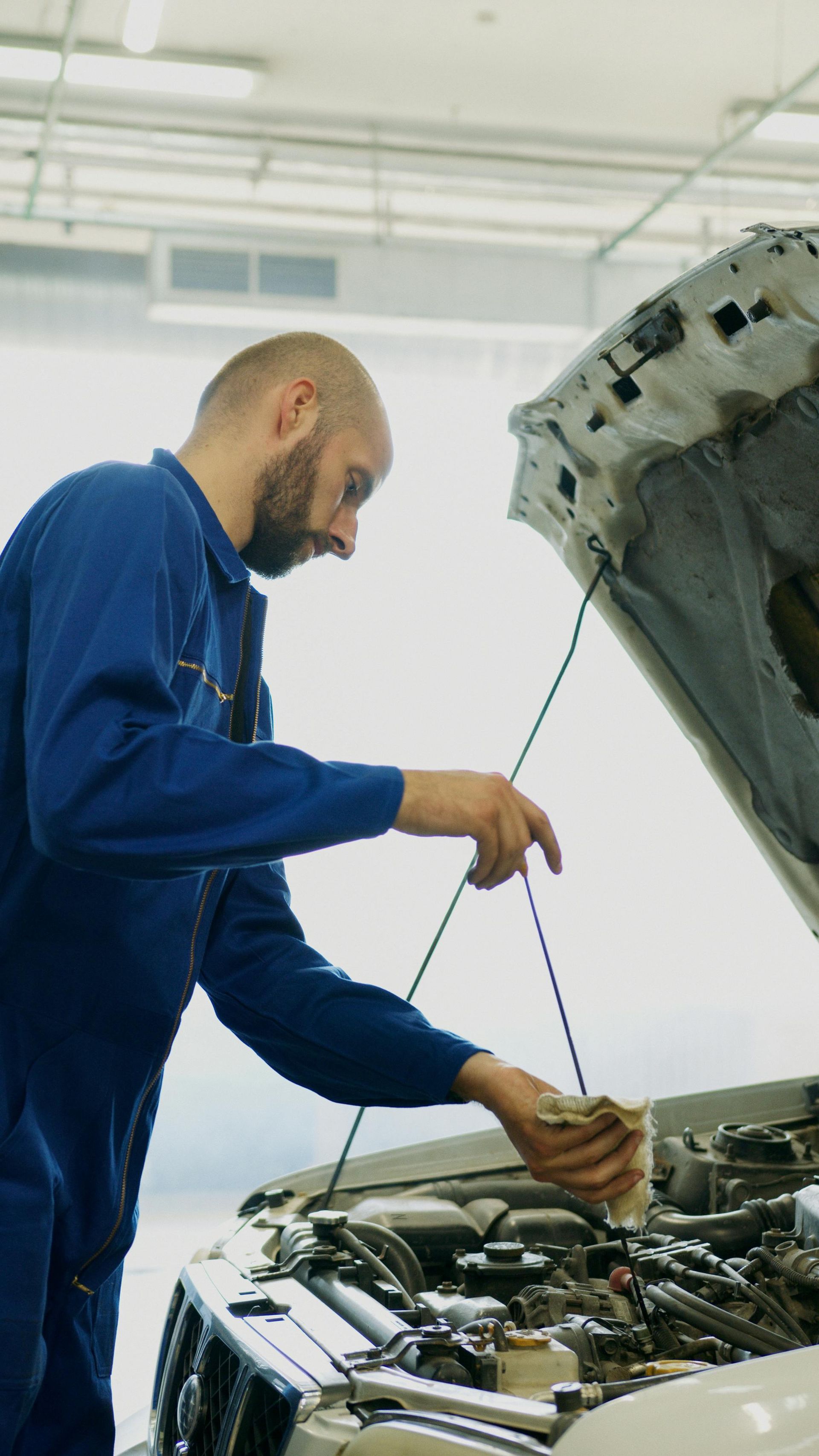 Mechanic in blue jumpsuit checks car engine, wiping with a cloth under open hood. Garage setting.