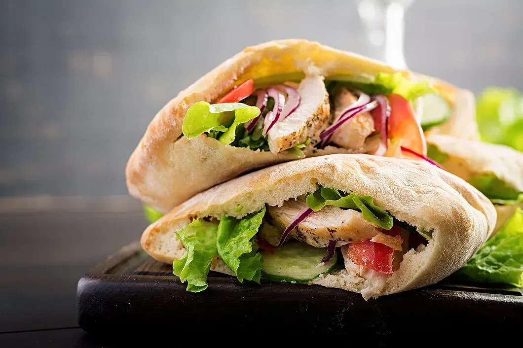 A close up of a pita sandwich with chicken and vegetables on a wooden cutting board.