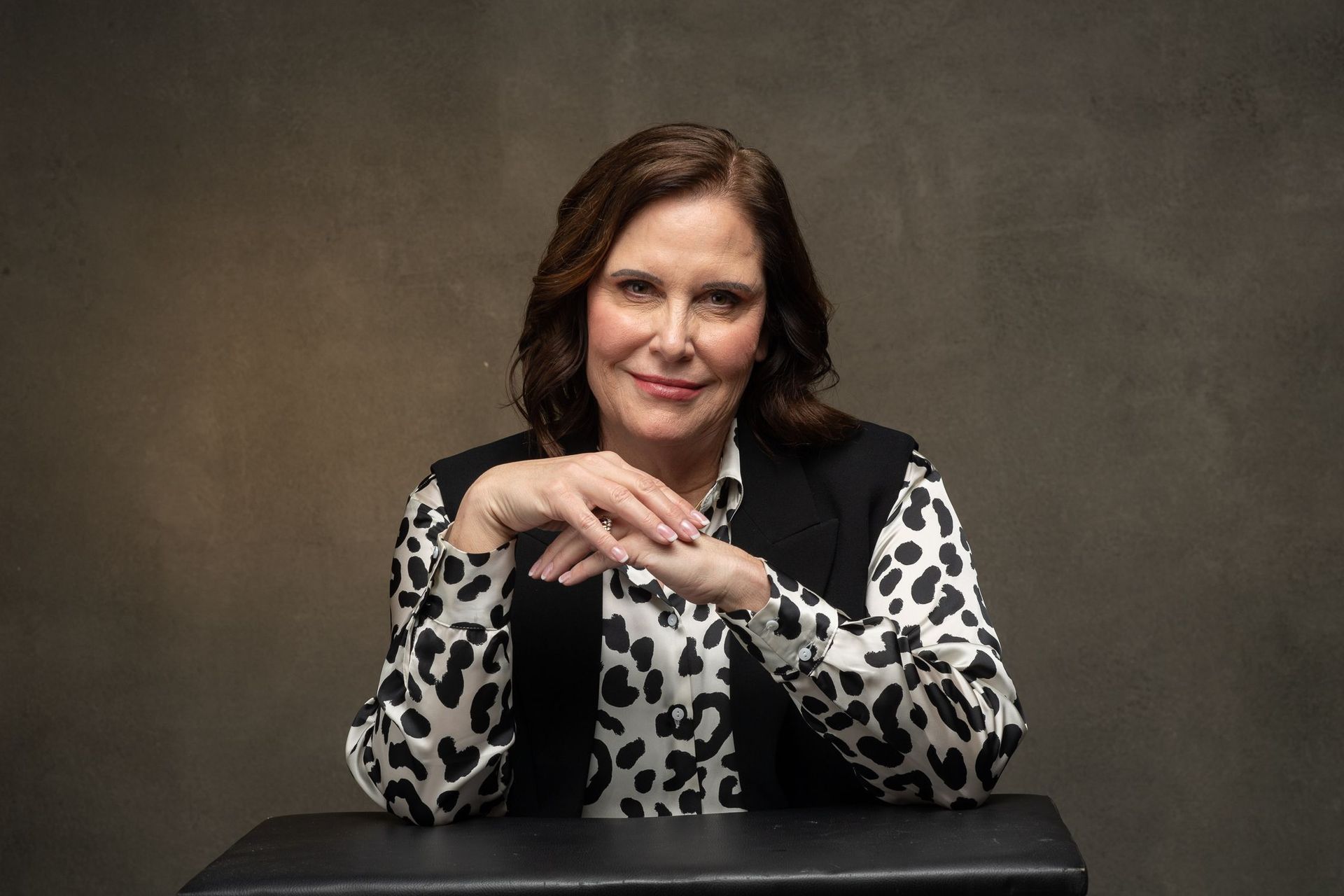 Woman seated with hands clasped, wearing black and white patterned shirt and vest, against a neutral background.