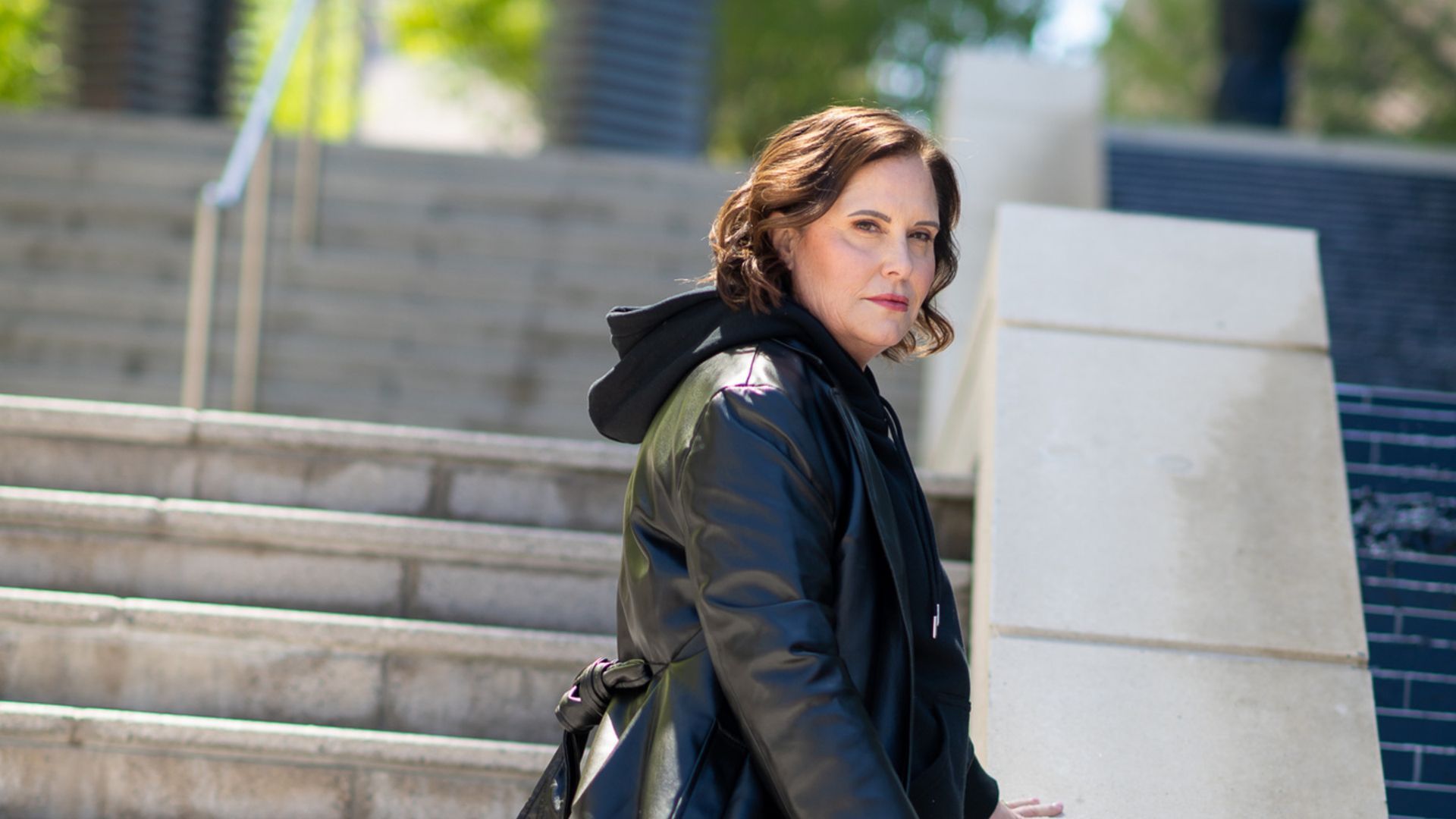 Woman in black jacket and hoodie on concrete stairs, looking over shoulder.