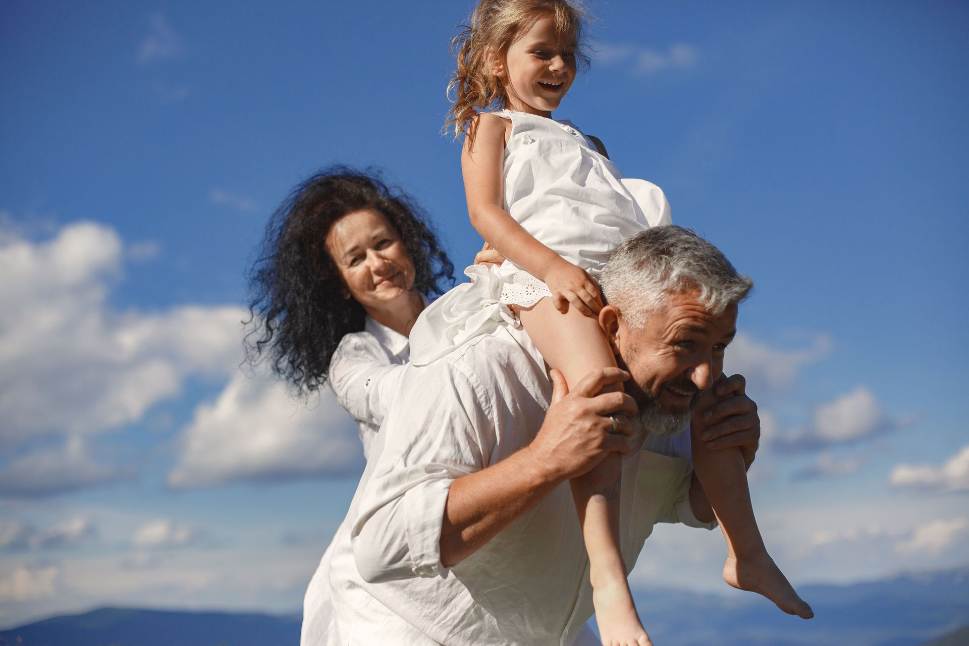 Two people giving a smiling child a piggyback ride outdoors under a blue sky
