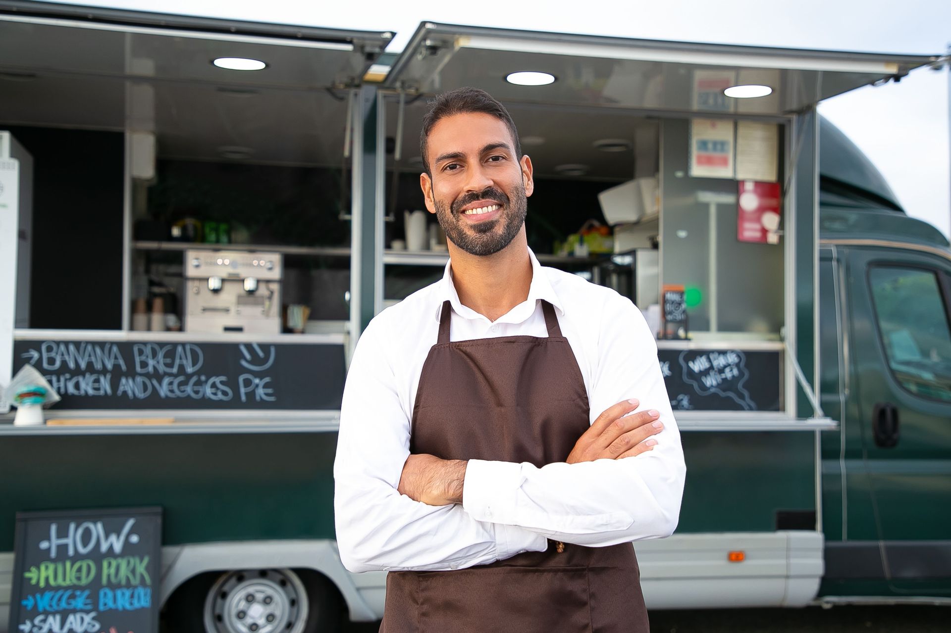 Smiling man in a brown apron stands in front of a food truck with arms crossed.