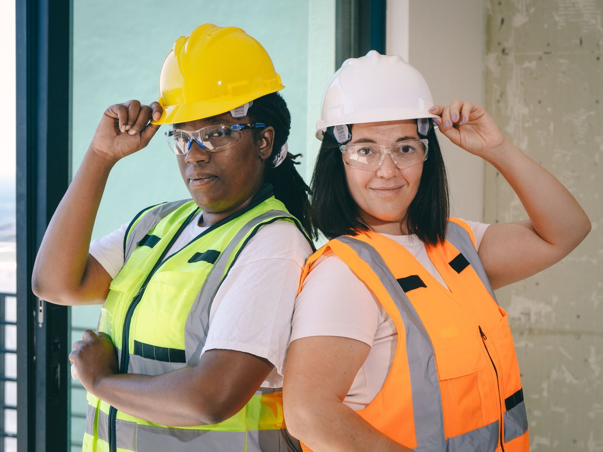 Two construction workers in hard hats and safety vests posing with hands on helmets.