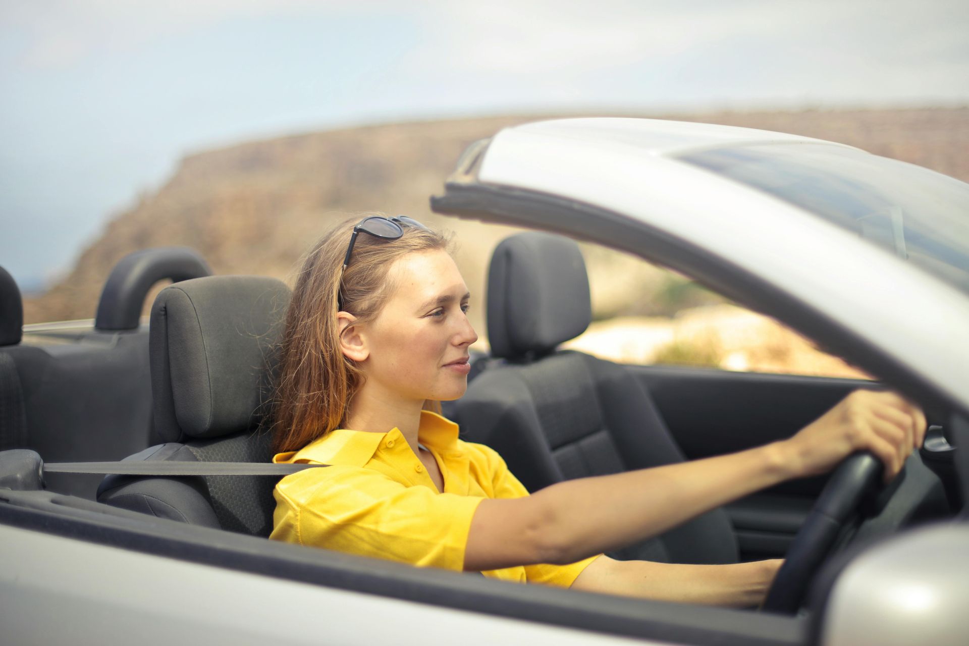 Woman driving a convertible, wearing a yellow shirt, with a blurred desert landscape of Abilene, Texas in the background