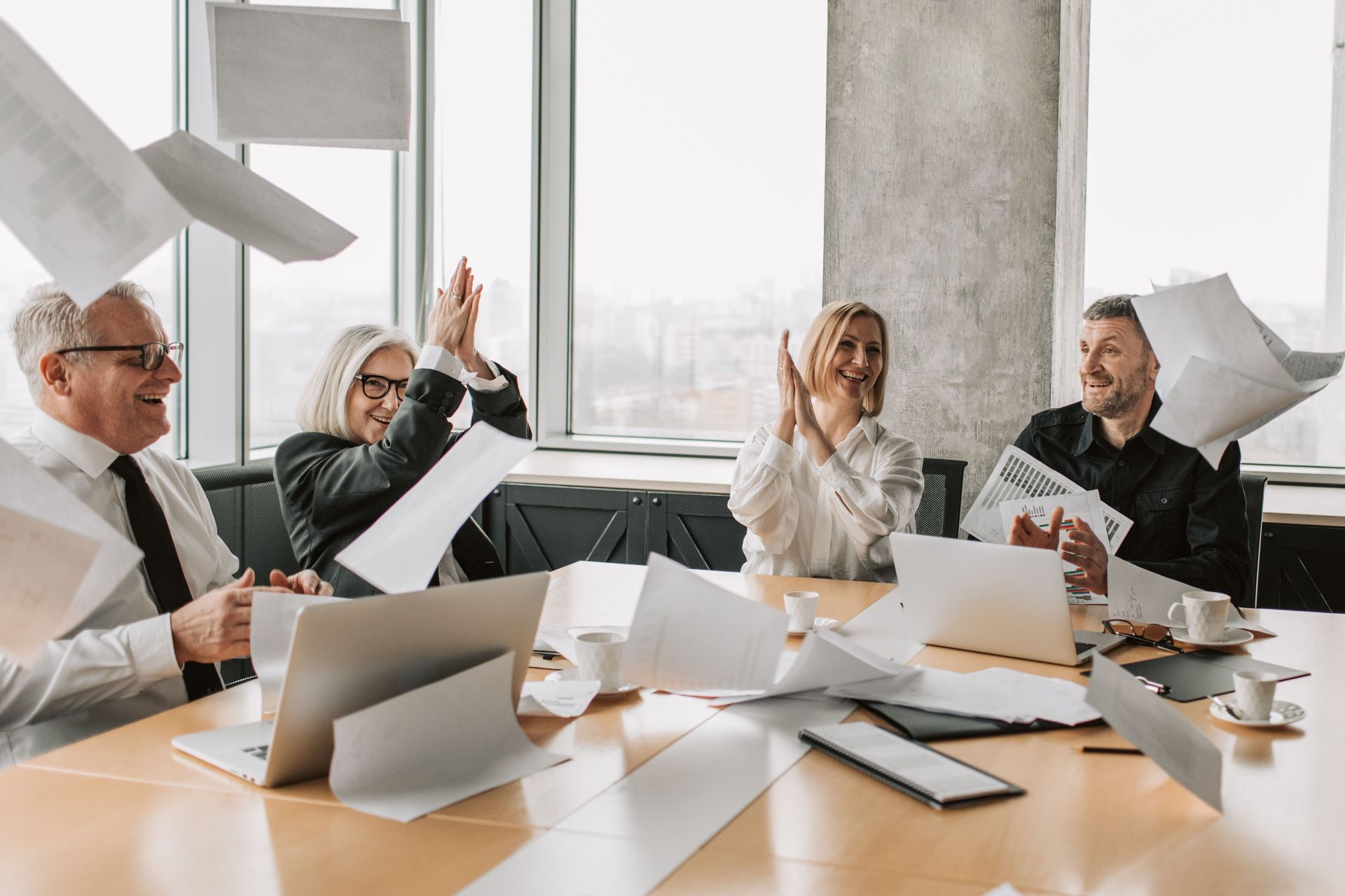 Team meeting in a bright office, coworkers around laptops with papers in the air, one person raising a hand.