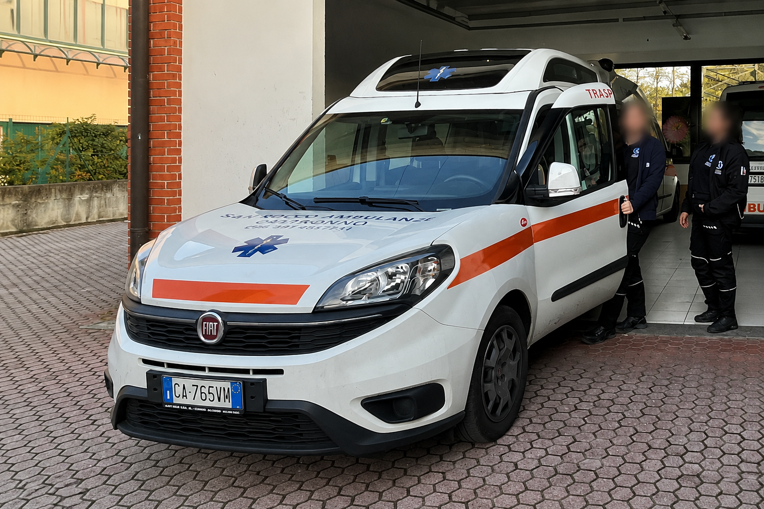 Veicolo di emergenza bianco con striscia arancione parcheggiato vicino a un edificio, con due persone in piedi accanto.