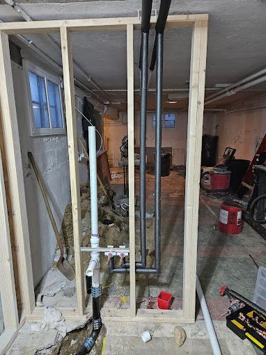 Unfinished basement interior featuring wood wall framing, exposed plumbing pipes with foam insulation, and a white drainpipe.