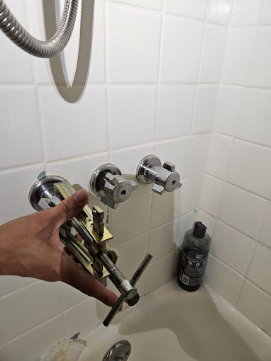 A hand holds a plumbing valve stem puller tool in front of a tiled shower wall with two installed faucet handles.