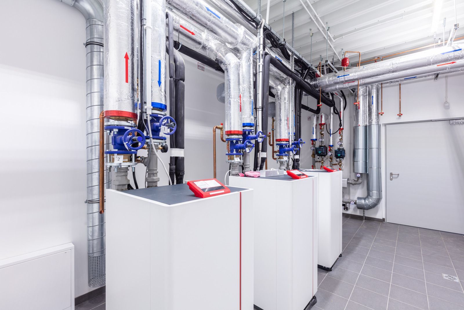 Three white heating units with silver insulated pipes and blue valves in a clean, utility room with gray tiled floors.