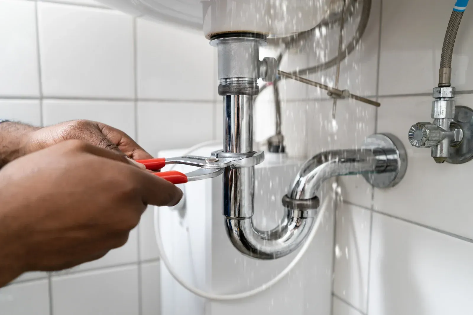 A person uses red-handled pliers to tighten a metal P-trap pipe under a bathroom sink against a white tiled wall.
