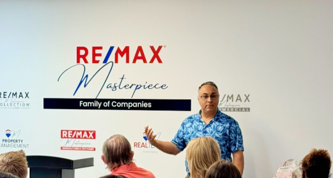A speaker in a blue patterned shirt gestures while presenting in front of a RE/MAX Masterpiece branded wall to an audience.