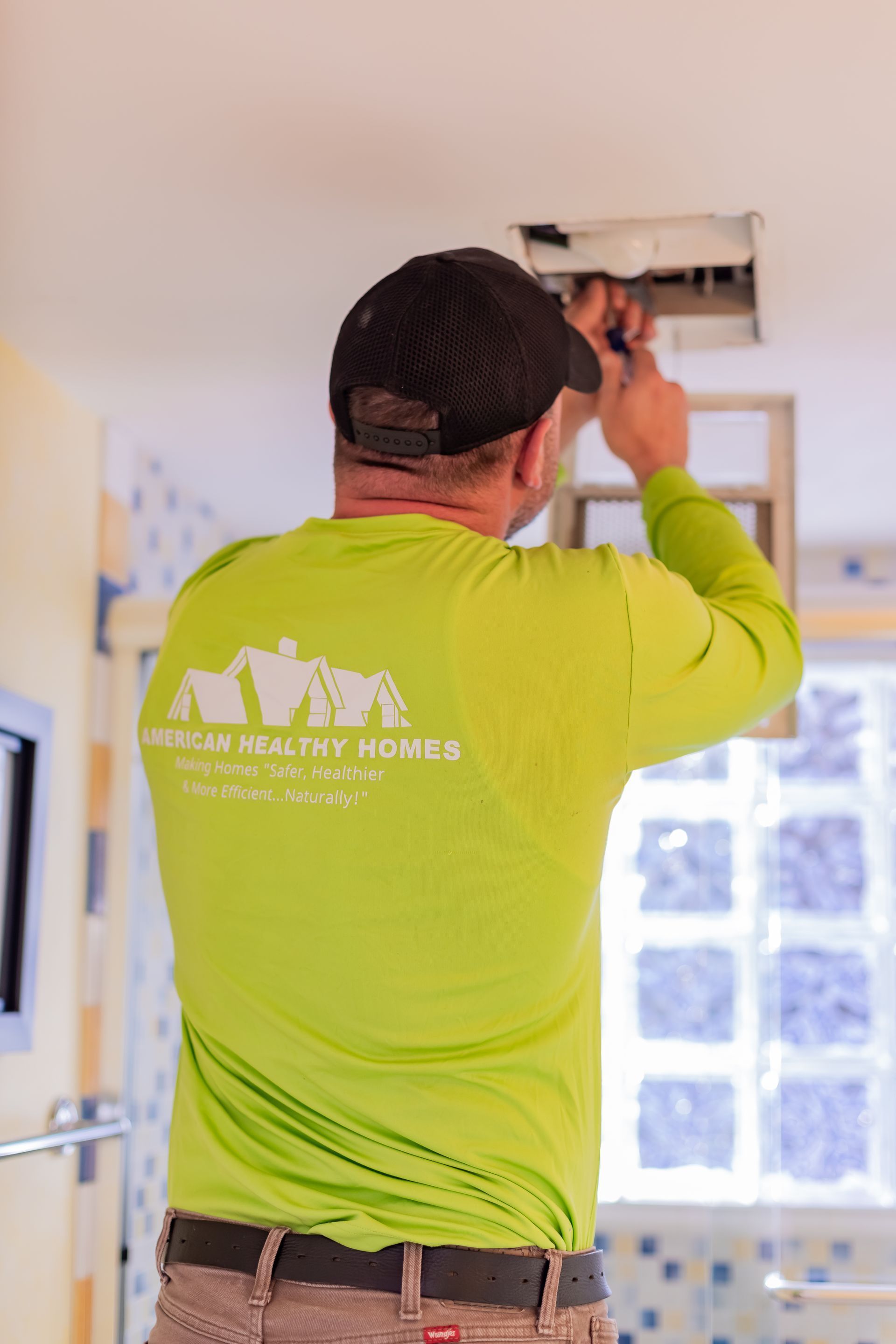 A worker in a lime-green long-sleeved shirt and black cap is installing or repairing a ceiling ventilation unit.