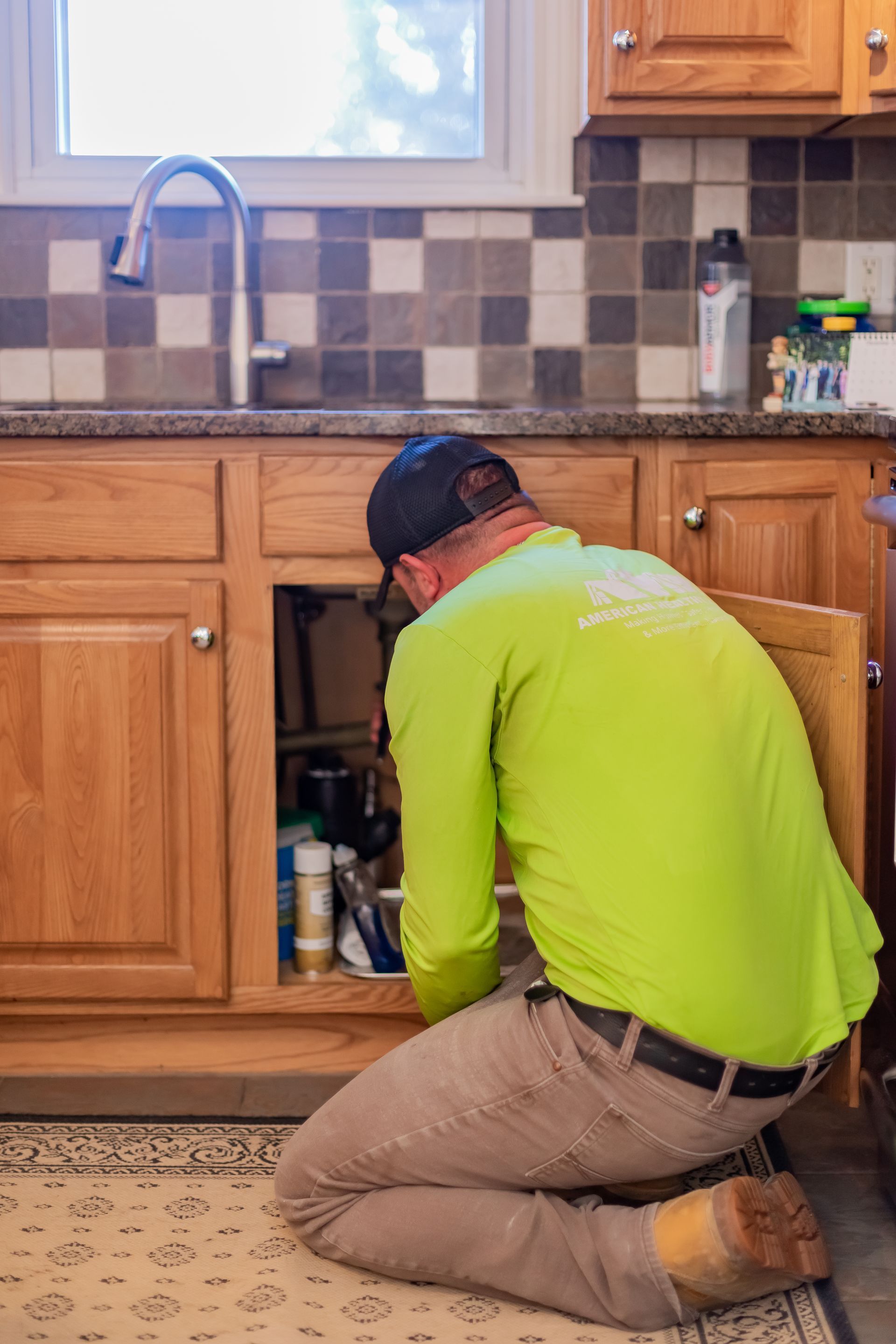 A worker in a neon green long-sleeve shirt kneels on a kitchen rug, repairing plumbing beneath a kitchen sink.