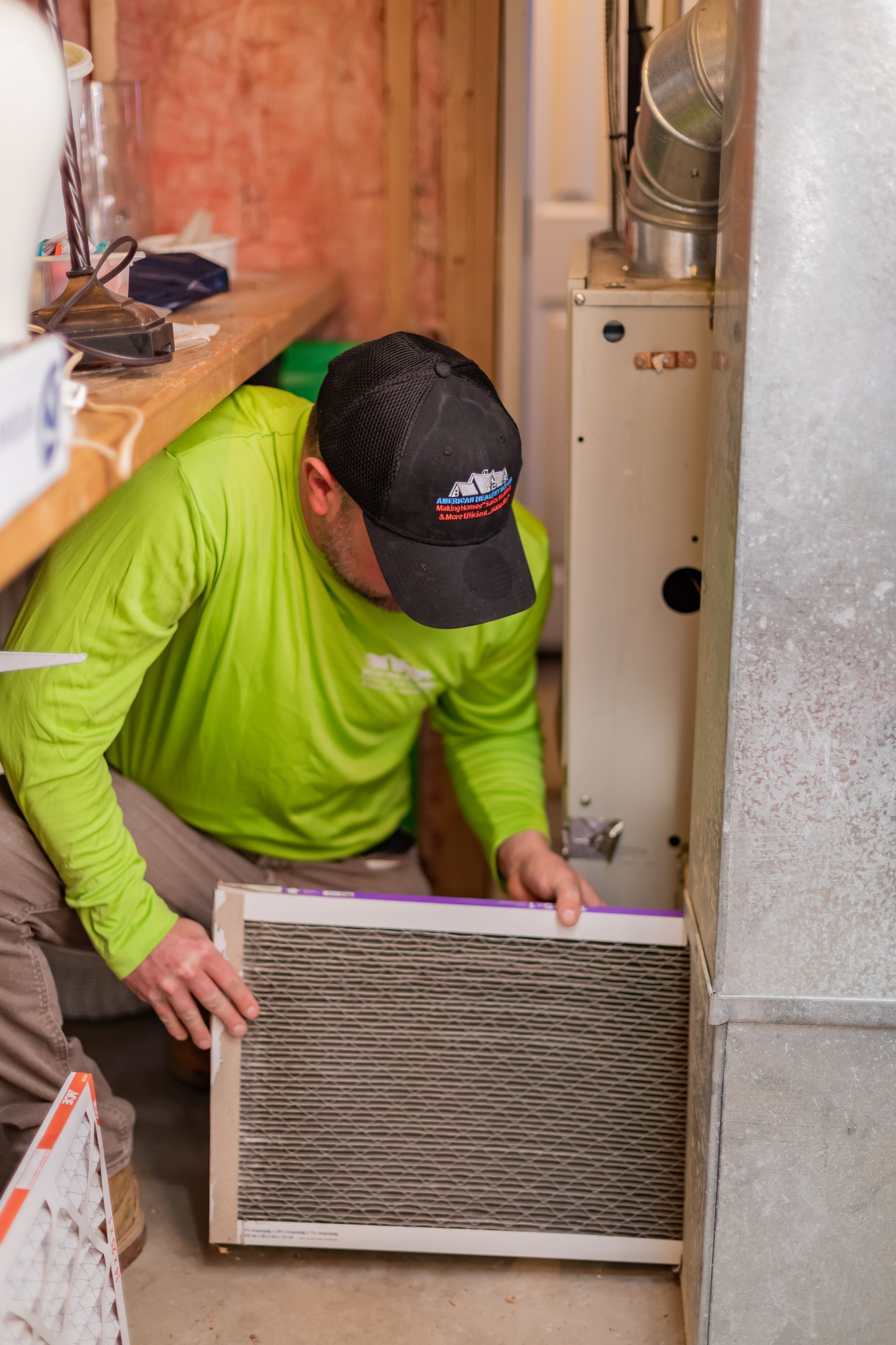 A technician in a lime green shirt replaces a dark air filter into a residential furnace system.