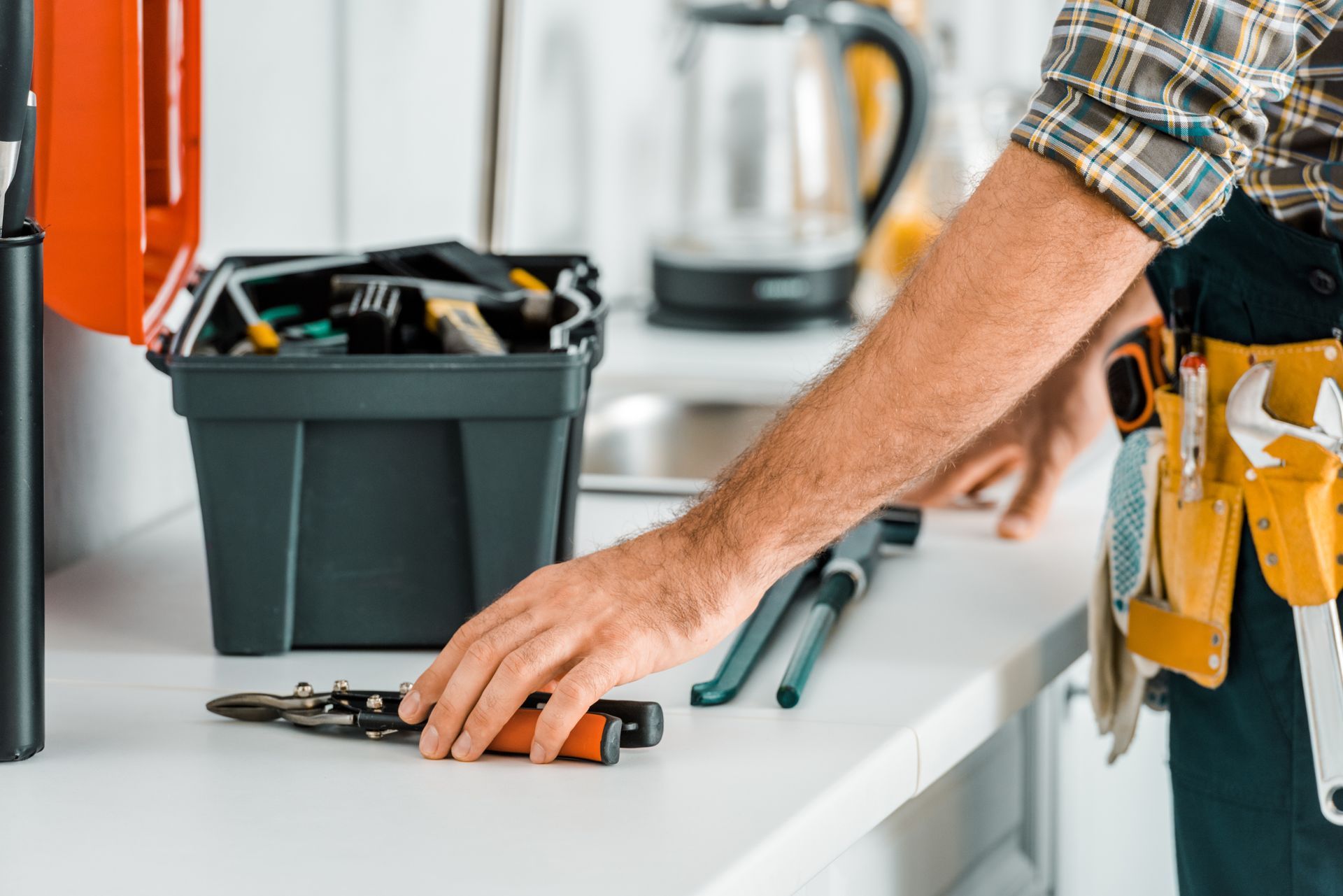 A person in work clothes and a tool belt prepares to pick up a tool from a white surface next to a black toolbox.