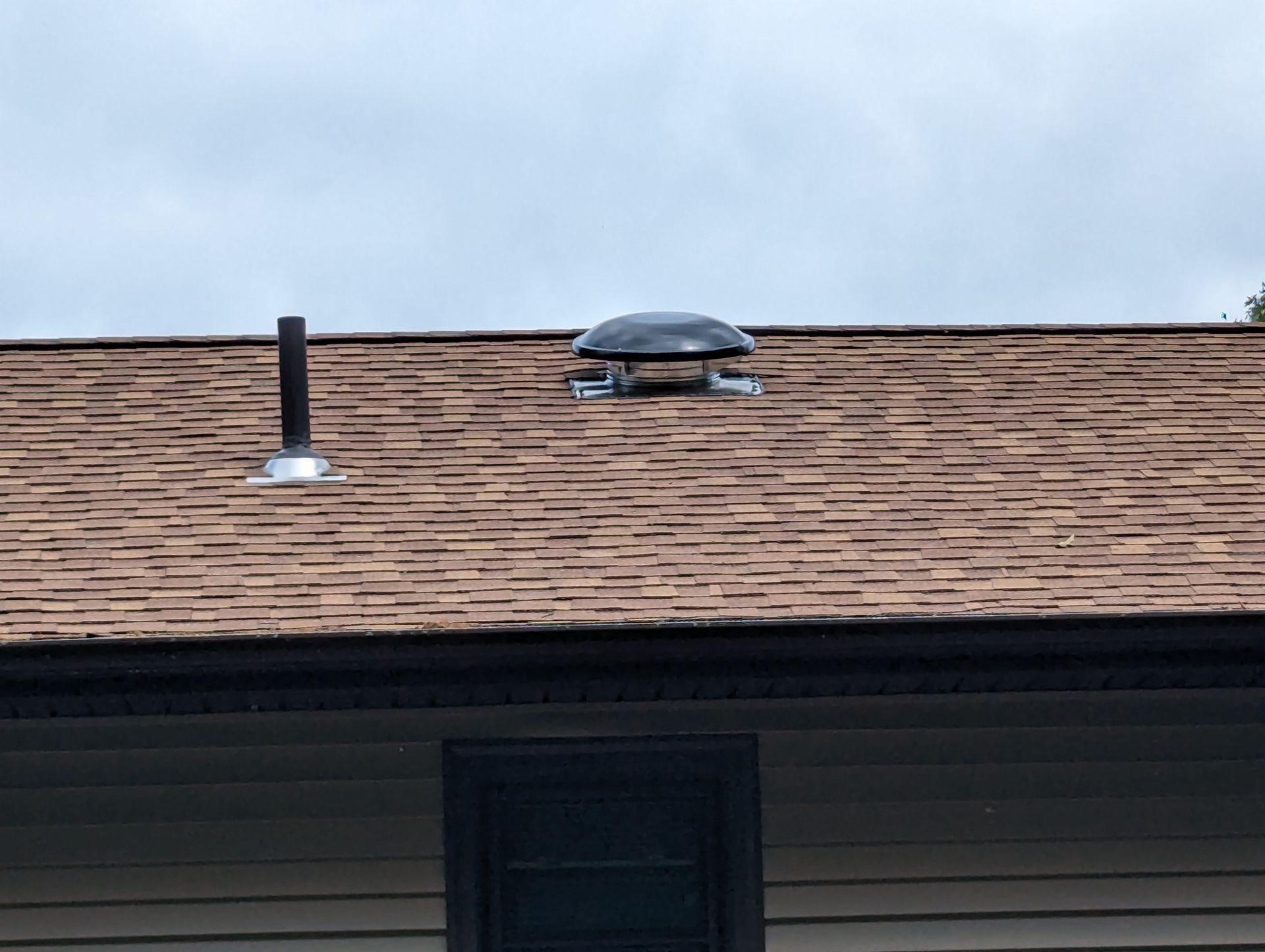 A brown asphalt-shingled roof featuring a plumbing vent pipe and a dome-shaped roof vent against a cloudy sky.