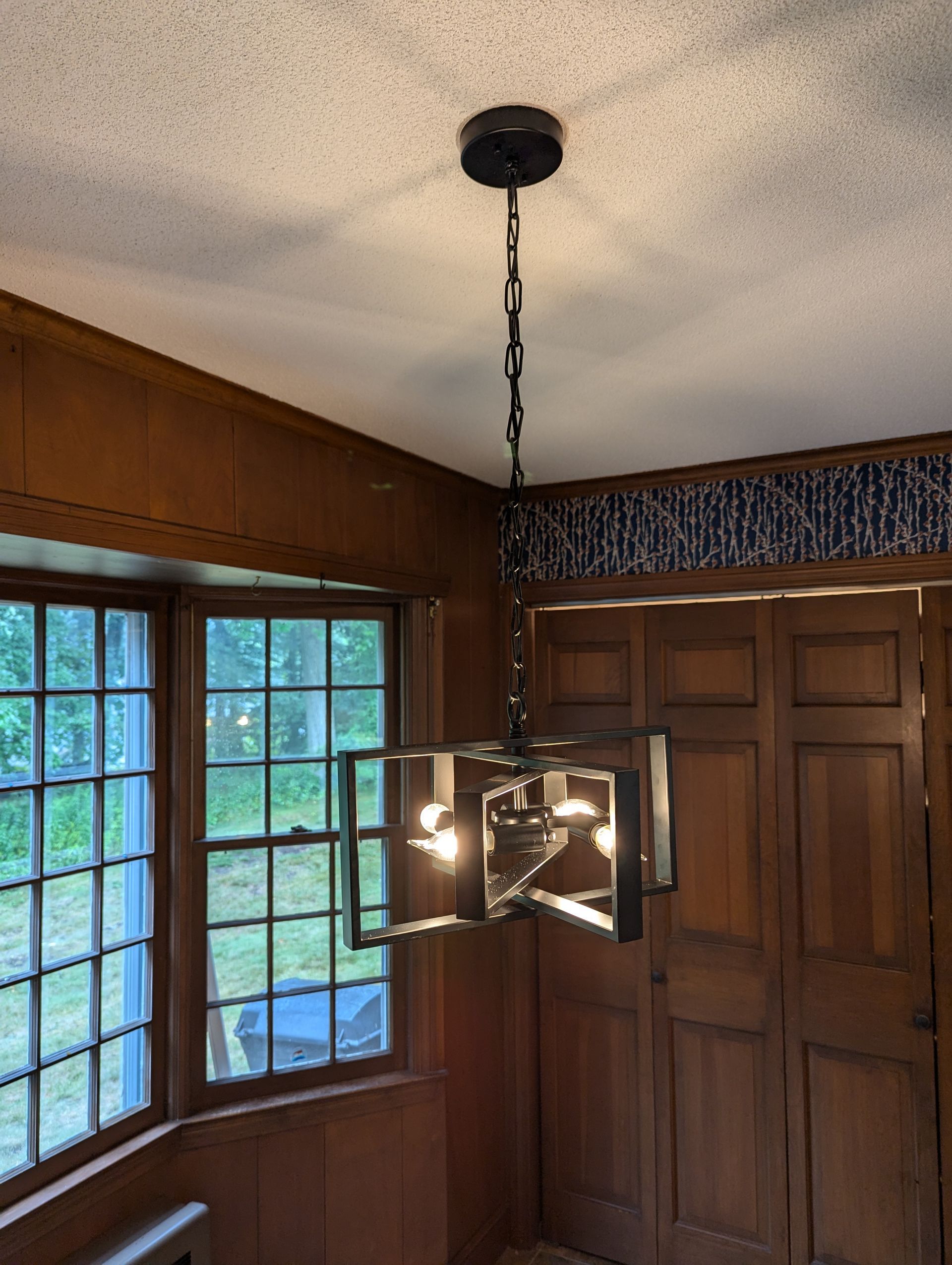 A modern, box-frame metal chandelier hangs from the ceiling in a room with wood-paneled walls and a window.
