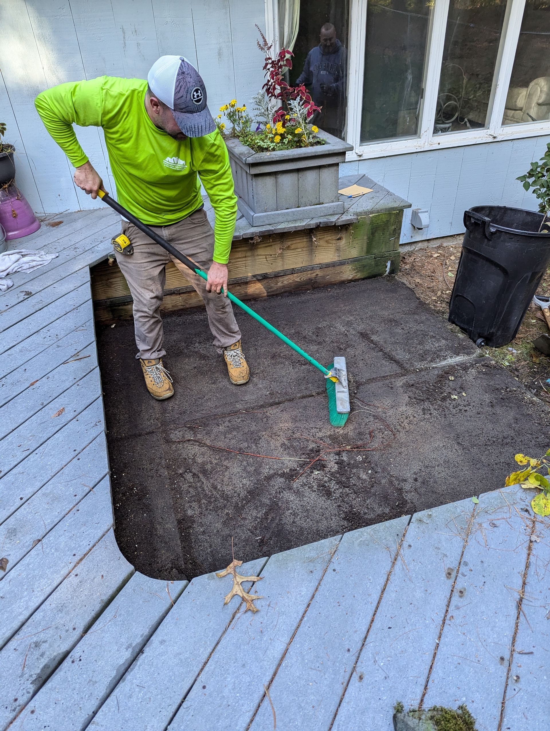 A person in a neon yellow long-sleeved shirt uses a long-handled landscaping tool to level soil in a cutout of a deck.