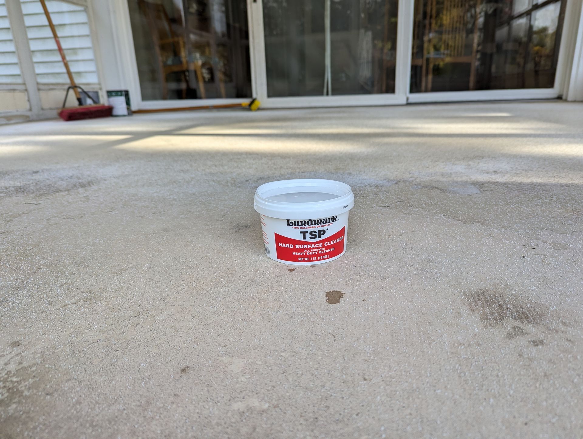 A small plastic container of TSP cleaner sits on a gray concrete patio in front of a glass sliding door.
