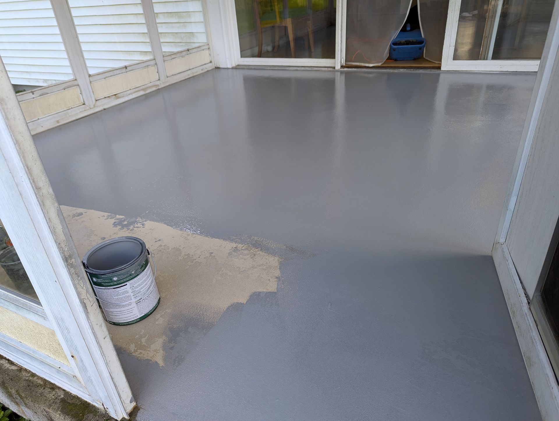 A partially painted gray floor on a covered porch, with a paint can sitting on an unfinished section of concrete.