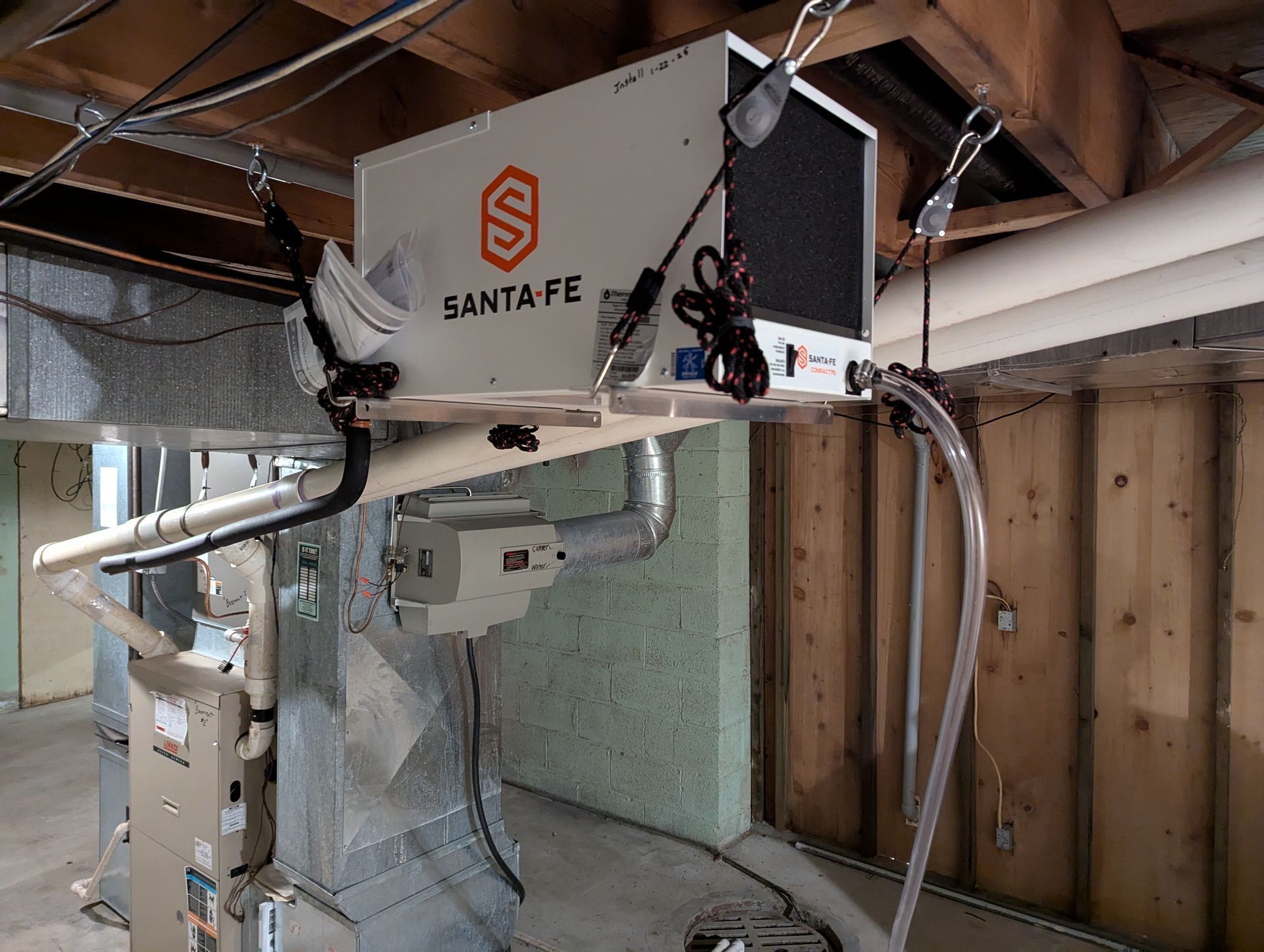 A white Santa Fe dehumidifier suspended from wooden ceiling joists above a basement furnace.