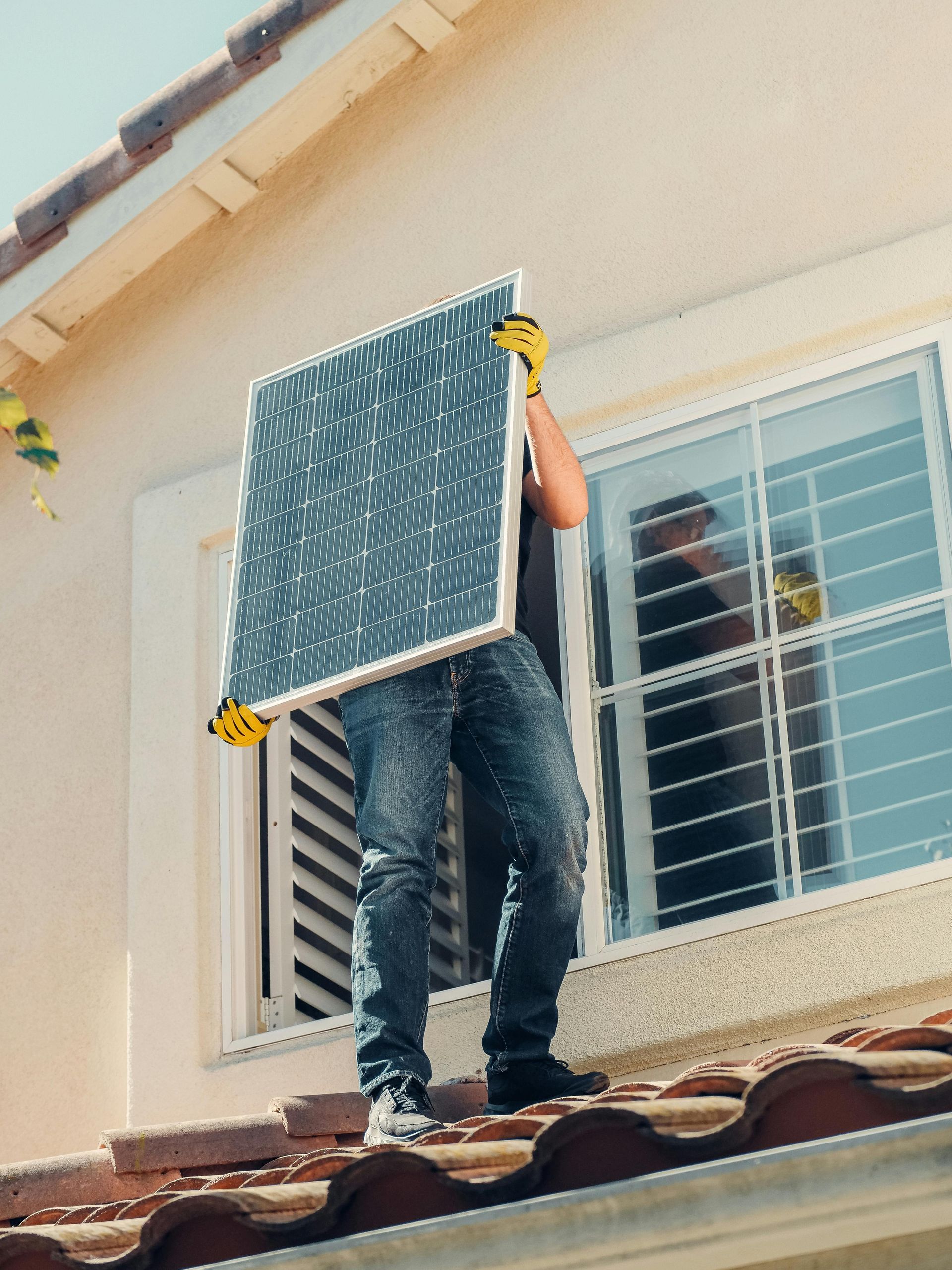Man carrying a solar panel on a rooftop. Sunny day. Beige house, terracotta roof tiles.