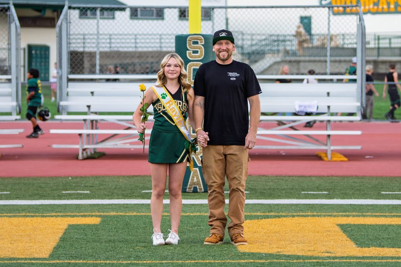 A young woman in a green cheerleading outfit and a man in a black shirt holding hands on a football field.