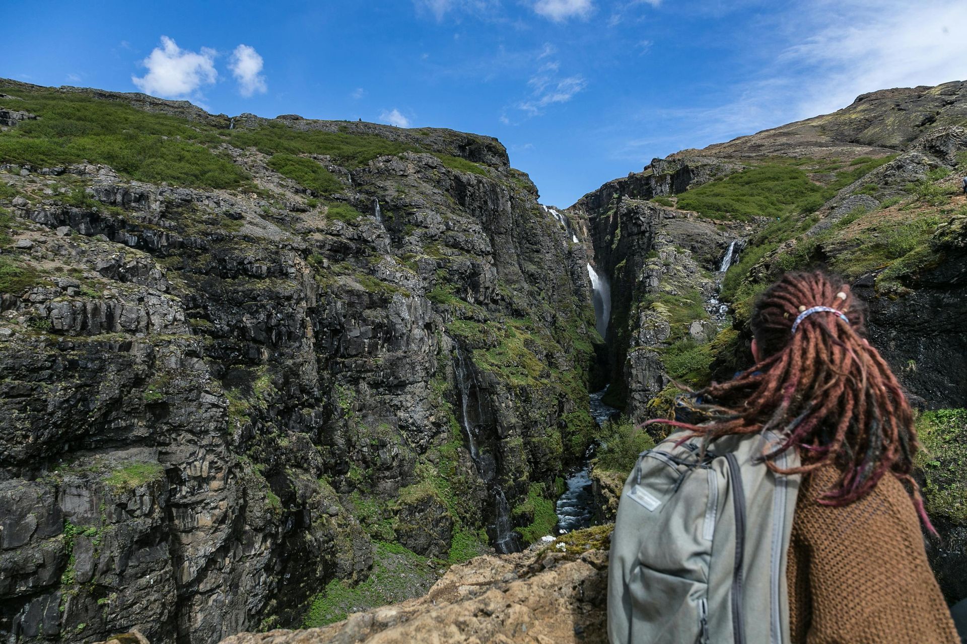 Person with dreadlocks, backpack overlooking a canyon and waterfall under a blue sky.