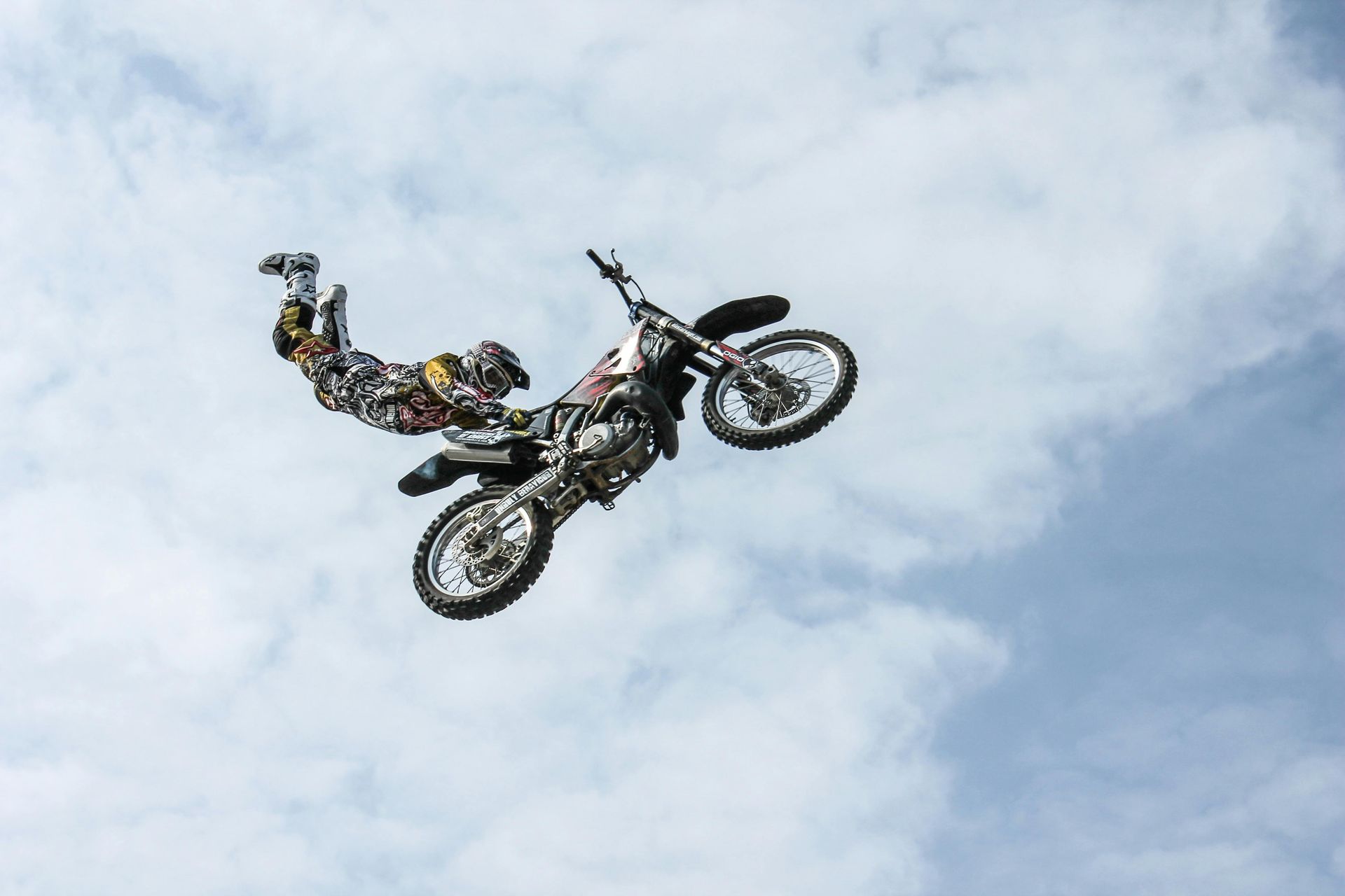 Motorcycle stunt rider airborne, performing a trick against a cloudy sky.