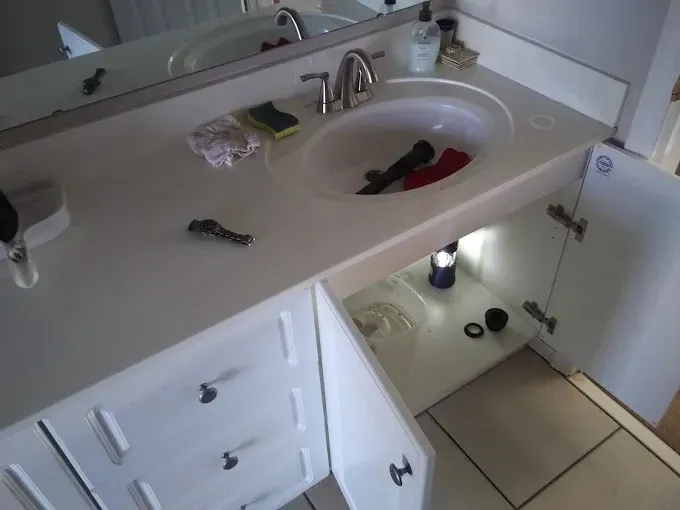 Bathroom sink cabinet with open doors, tools in the sink, and a flashlight inside.